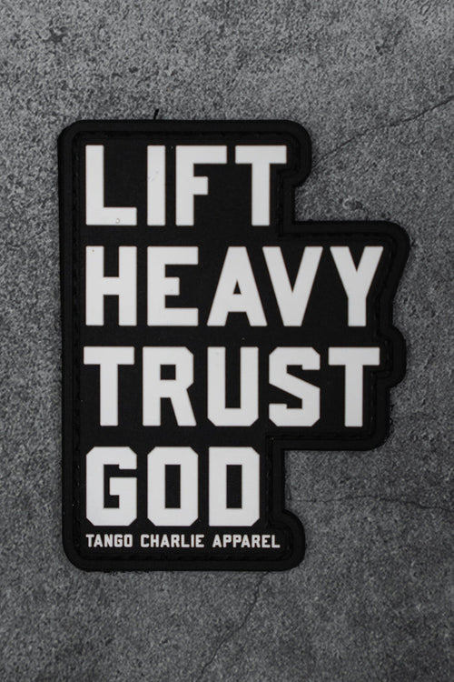 The Lift Heavy. Trust God. - Patch by Tango Charlie Apparel is a black 3D PVC patch with bold white text, shown on a gray textured background.