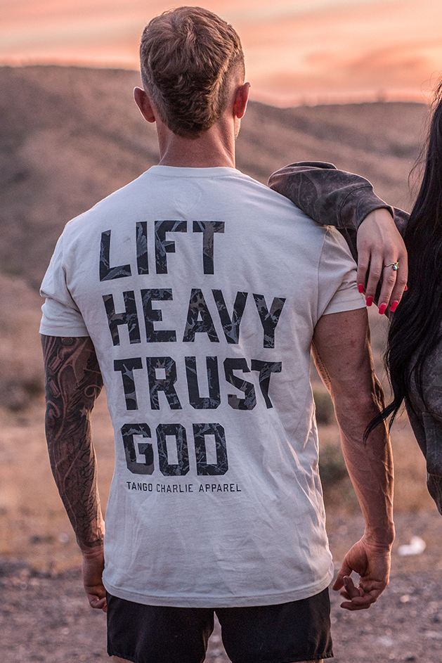 A man stands outdoors with his back to the camera, wearing the Tango Charlie Apparel "Lift Heavy. Trust God. - Tee," inspired by Christian fitness and Proverbs 3:5-6. A woman stands beside him, her arm on his shoulder as dry hills stretch out behind them.