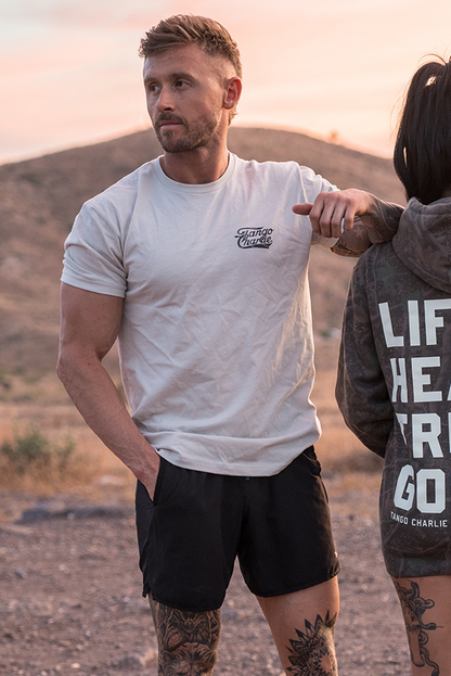 A man wears the "Lift Heavy. Trust God." tee by Tango Charlie Apparel, standing outdoors with one hand in his pocket and the other on a woman's shoulder, hills and a sunset sky highlighting this Christian fitness scene.