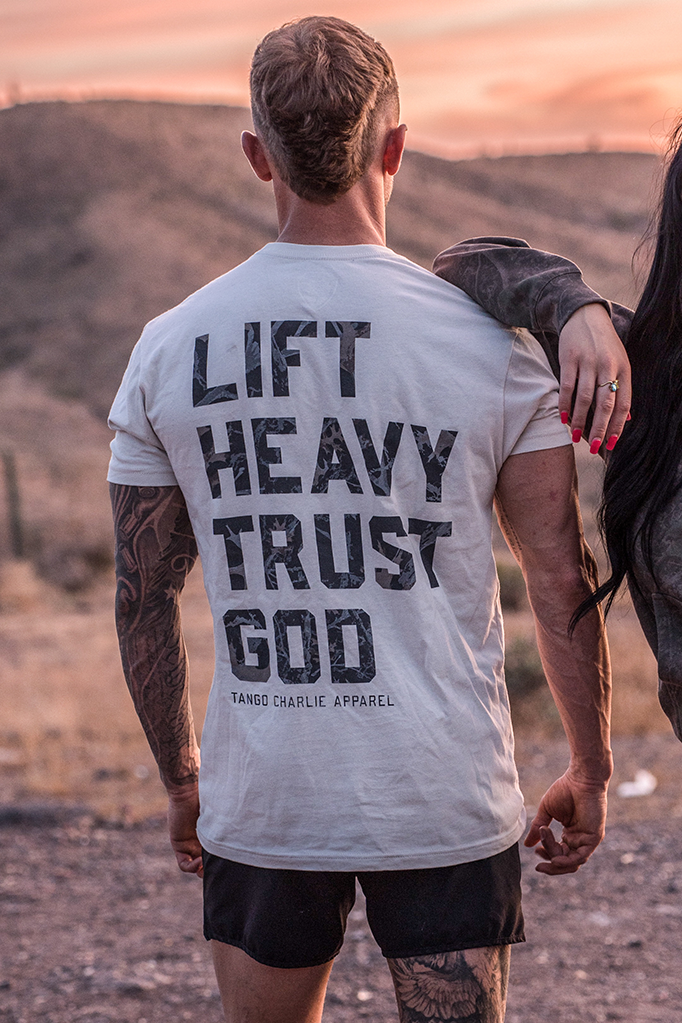 A man faces away from the camera, wearing Tango Charlie Apparel’s “Lift Heavy. Trust God. - Tee.” Another person rests their arm on his shoulder as sunset hills glow, reflecting Christian fitness and the spirit of Proverbs 3:5-6.