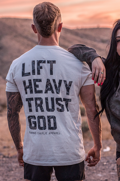 A person outdoors wears the "Lift Heavy. Trust God. - Tee" by Tango Charlie Apparel, featuring bold black text on a white shirt, embracing Christian fitness as the sunset glows behind them over rolling hills.