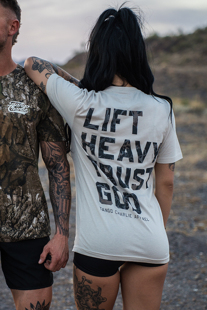 A woman with long dark hair in a ponytail wears the "Lift Heavy. Trust God. - Tee" by Tango Charlie Apparel, standing beside a tattooed man in camo. Smiling outdoors on a rocky path, they embody Christian fitness inspired by Proverbs 3:5-6.