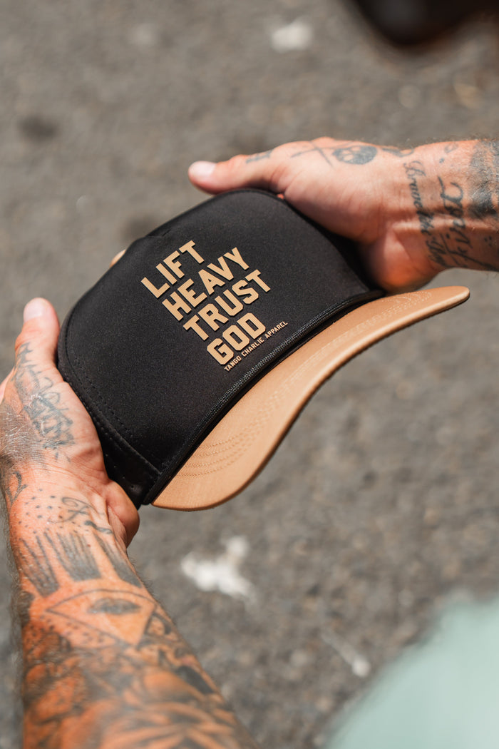 A tattooed individual holds the Lift Heavy. Trust God. Hydro - Black Snapback Hat by Tango Charlie Apparel—a Christian gym hat crafted for both style and purpose, featuring the bold phrase “LIFT HEAVY. TRUST GOD.” across the front. This sweat-resistant snapback is ideal for anyone seeking motivation and faith during their toughest workouts.