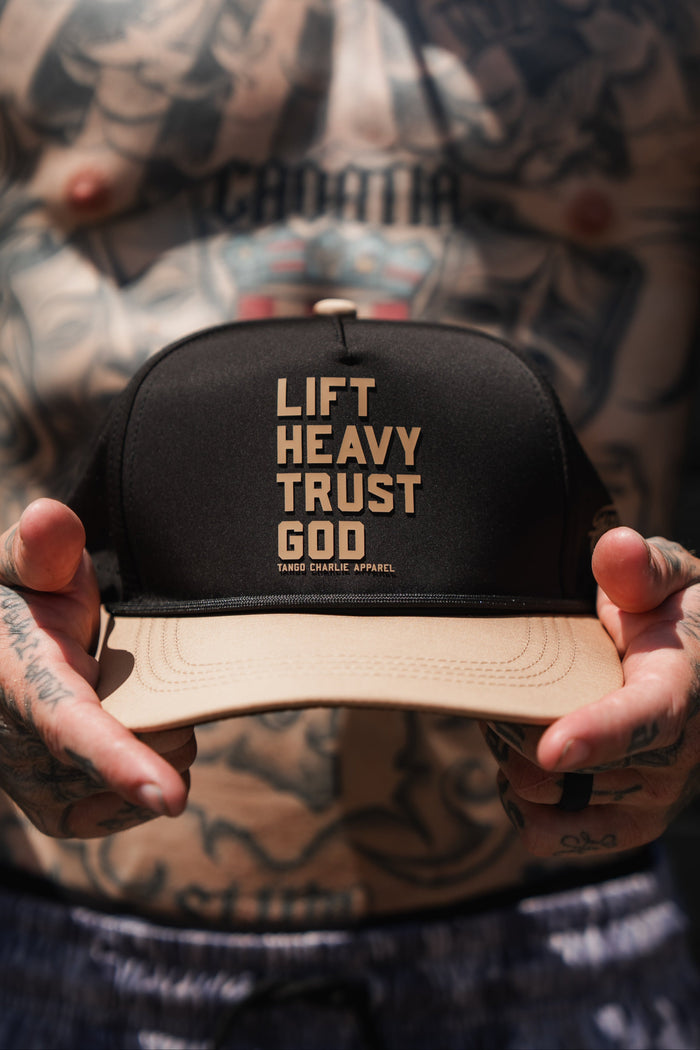 A tattooed person holds the Lift Heavy. Trust God. Hydro - Black Snapback Hat by Tango Charlie Apparel, a Christian gym hat that features LIFT HEAVY TRUST GOD in bold lettering on the front panel.