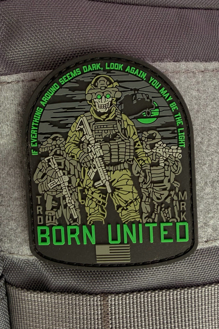 The Light in the Darkness PVC Patch by Born United Private Label features three armed soldiers with night vision goggles, a helicopter backdrop, and green text: “If everything around seems dark, look again, you may be the light. Born United.”.