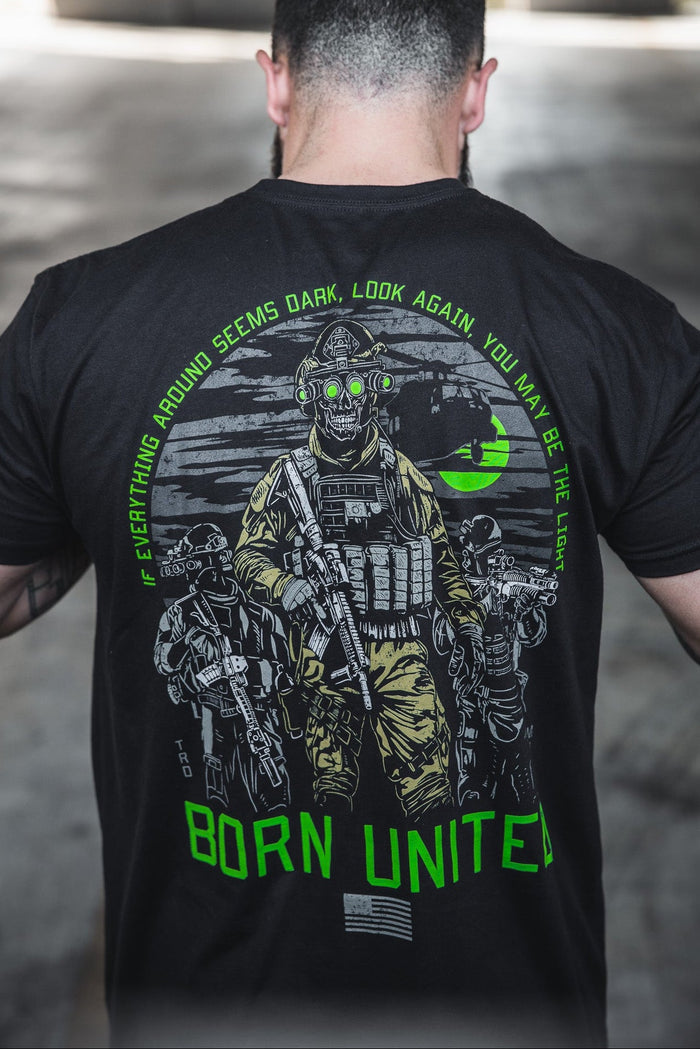 A man wears the Born United Private Label "Light in the Darkness" T-shirt, featuring armed soldiers with night vision goggles, a green moon, and the words: Everything around seems dark, look again—you may be the light.