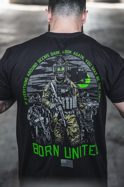 A man wears the Born United Private Label "Light in the Darkness" T-shirt, featuring armed soldiers with night vision goggles, a green moon, and the words: Everything around seems dark, look again—you may be the light.