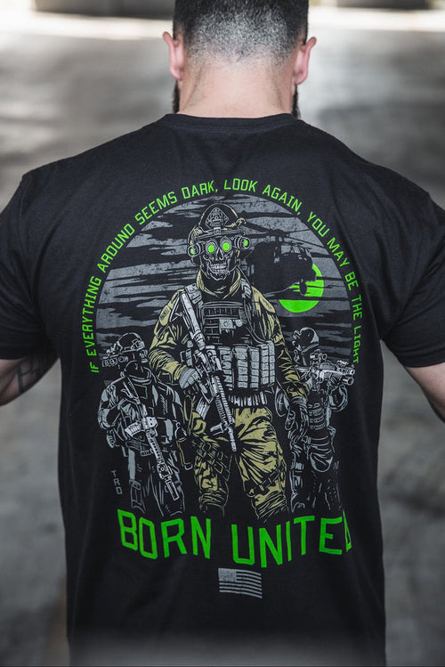 A man wears the Born United Private Label "Light in the Darkness" T-shirt, featuring armed soldiers with night vision goggles, a green moon, and the words: Everything around seems dark, look again—you may be the light.