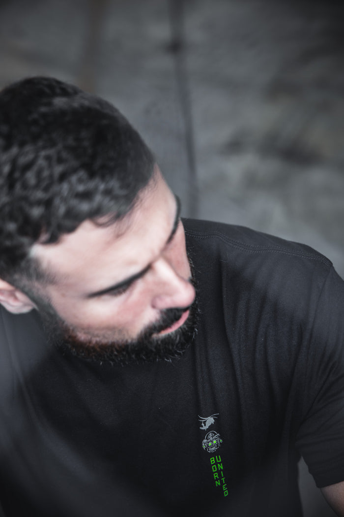 A man with dark hair and a beard looks down, wearing Born United Private Label's "Light in the Darkness" black t-shirt. The blurred background features a soft, moody effect with subtle 60/40 color blending.