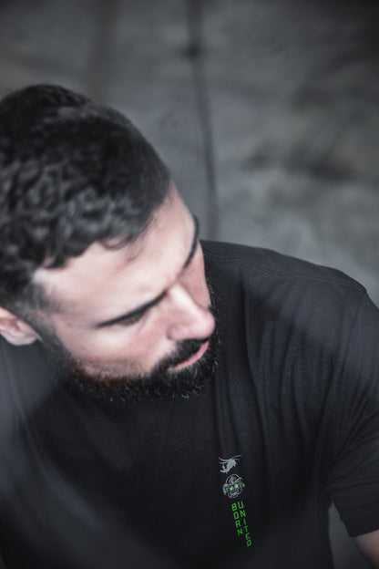 A man with dark hair and a beard looks down, wearing Born United Private Label's "Light in the Darkness" black t-shirt. The blurred background features a soft, moody effect with subtle 60/40 color blending.