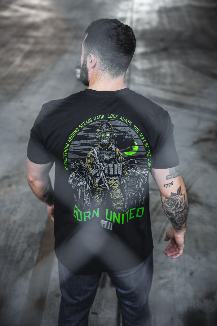 A man faces away from the camera wearing the Born United Private Label’s "Light in the Darkness" black t-shirt featuring a soldier with night vision goggles and motivational text: "BORN UNITED" and "If everything around seems dark...you may be the Light.