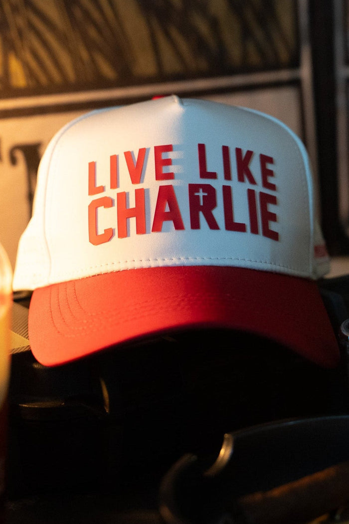 The Born United Private Label Live Like Charlie Ivory Snapback, featuring "LIVE LIKE CHARLIE" and a small cross on the front, rests on a dark surface amid various objects in dim lighting.