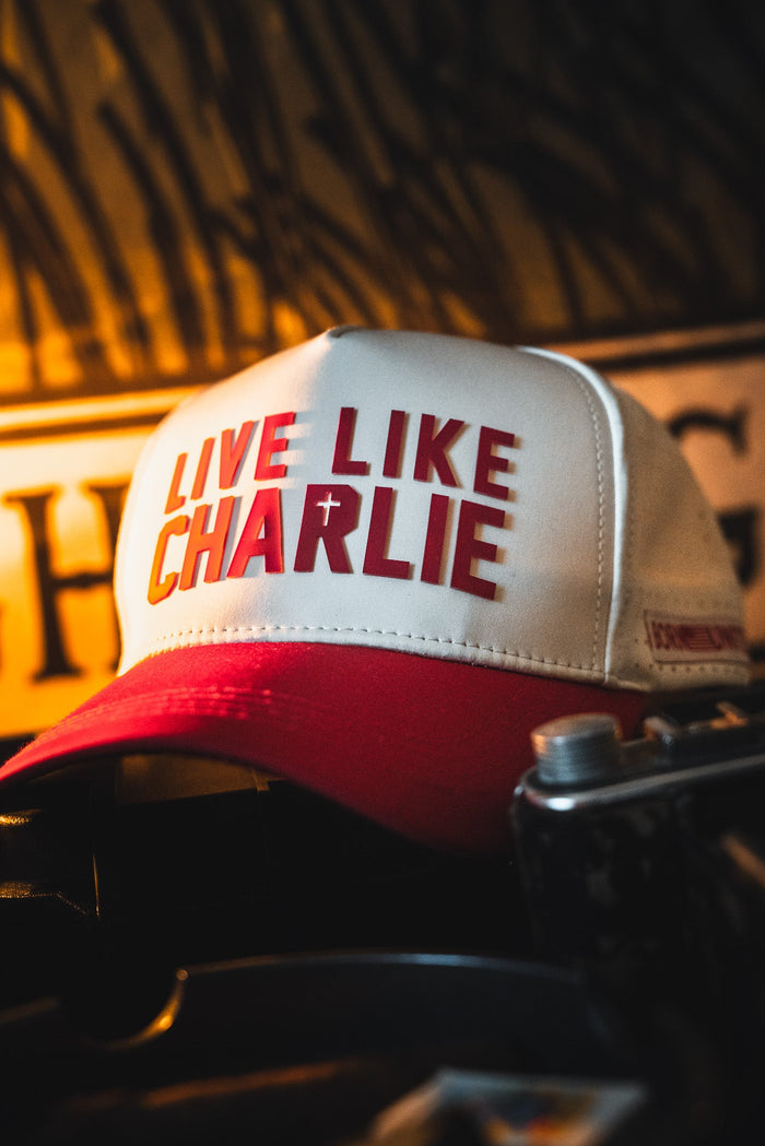 The Born United Private Label Live Like Charlie Ivory Snapback, featuring bold red lettering, is stylishly displayed against a warm, blurred background for a fashionable and comfortable look.