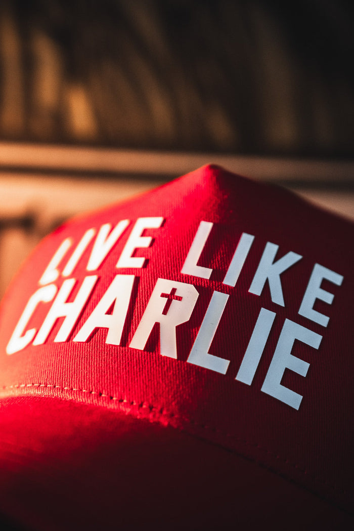 The Live Like Charlie Trucker Snapback by Born United Private Label is a red cap featuring bold white "LIVE LIKE CHARLIE" text, with the "I" in "CHARLIE" as a cross, and a classic curved brim.