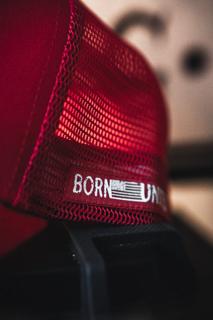 Close-up of a red mesh Live Like Charlie Trucker Snapback by Born United Private Label with a curved brim, featuring white "BORN U" text and a stylized American flag on the front. The background is blurred.