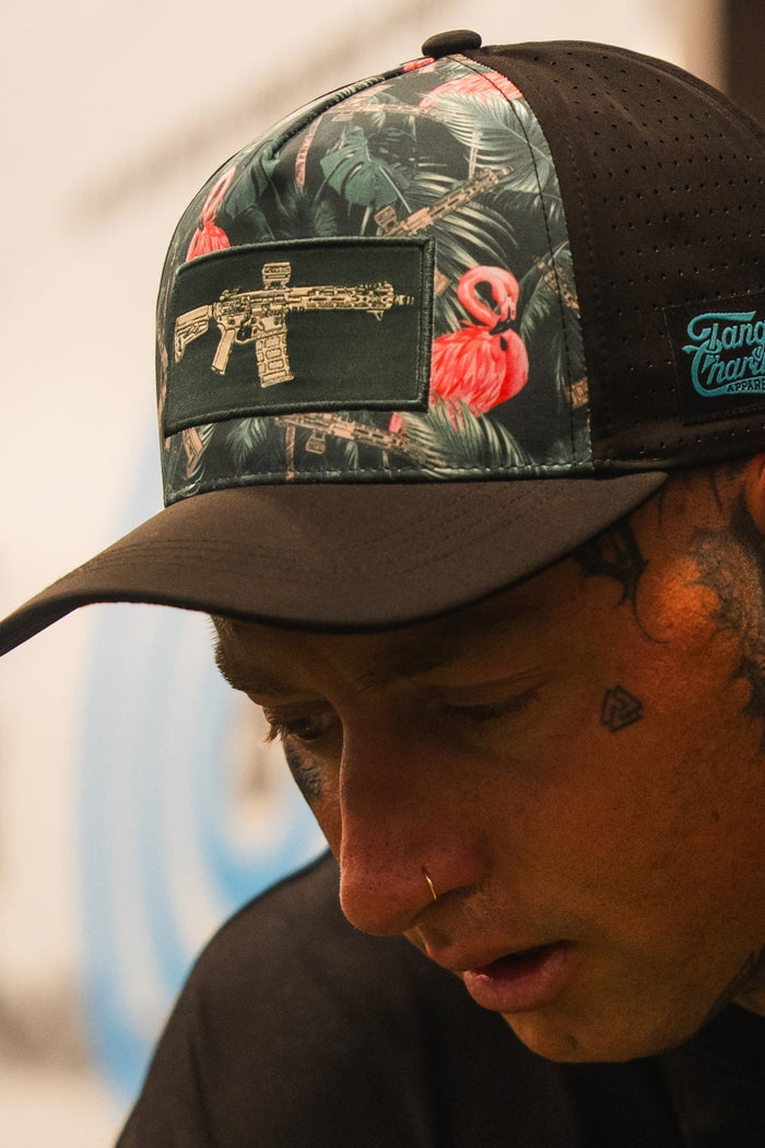 A person with face tattoos wears the Loaded Flamingo Hydro Snapback Hat by Tango Charlie Apparel, which has a black and floral design with a gold gun patch. They are looking down, with a blurred background behind them.