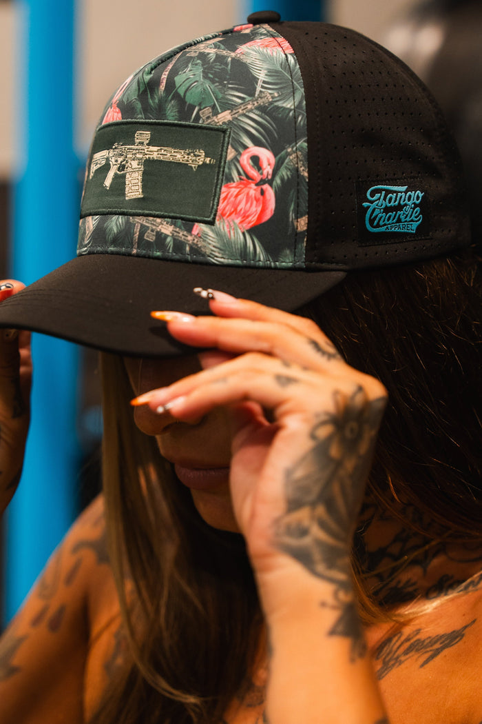 A tattooed woman adjusts a Loaded Flamingo Hydro Snapback Hat by Tango Charlie Apparel, featuring a floral flamingo design, submachine gun patch, and blue Tango Charlie logo. Her face is partially hidden.