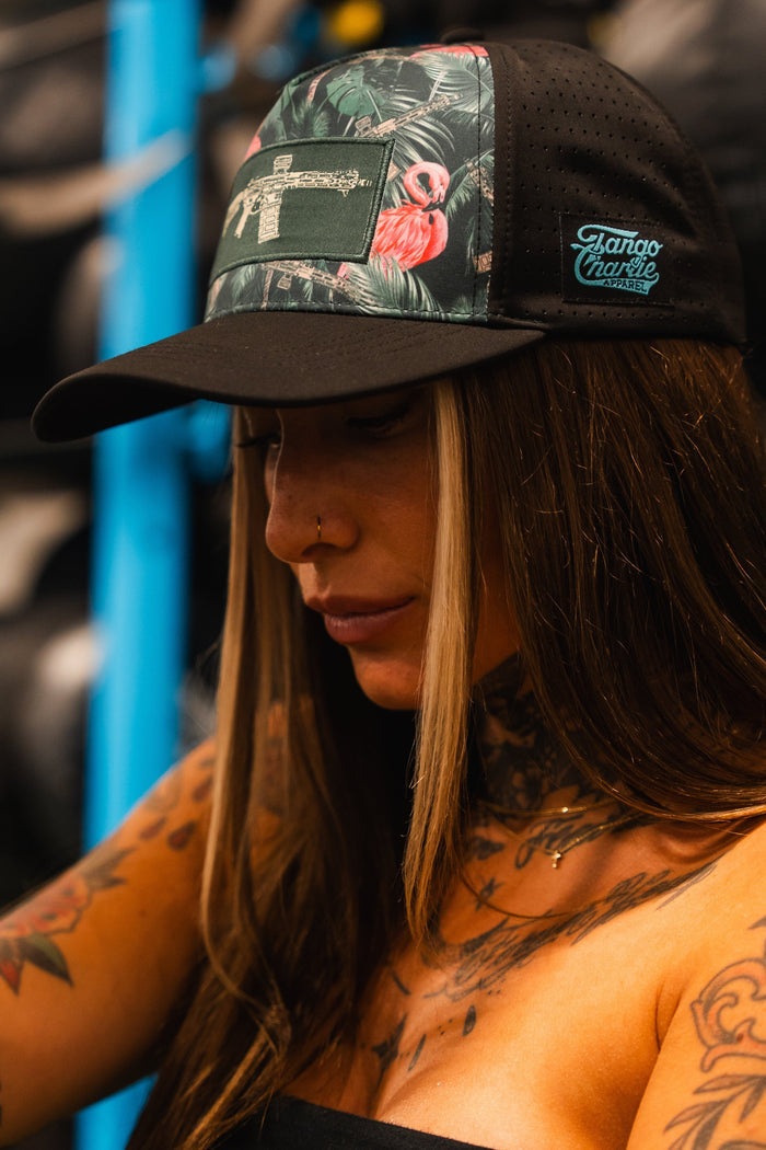A person with long brown hair and tattooed arms wears the Loaded Flamingo Hydro Snapback Hat by Tango Charlie Apparel, featuring a black and green tropical print with a gun design, standing indoors near gym equipment.