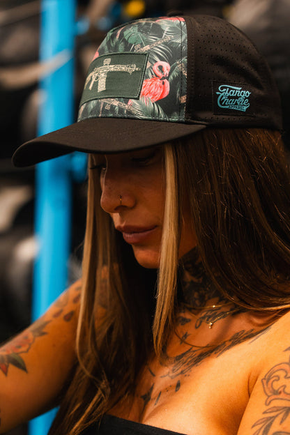 A person with long brown hair and tattooed arms wears the Loaded Flamingo Hydro Snapback Hat by Tango Charlie Apparel, featuring a black and green tropical print with a gun design, standing indoors near gym equipment.