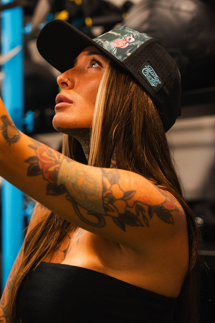 A woman with long hair and colorful arm tattoos wears the Loaded Flamingo Hydro Snapback Hat by Tango Charlie Apparel and a strapless top, looking upward and gripping a thick rope in a gym setting.