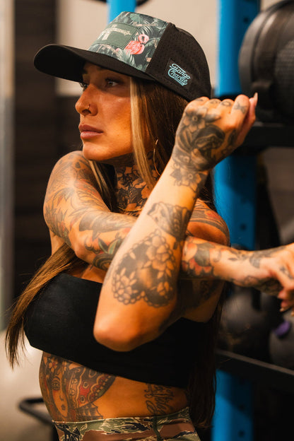 A tattooed woman wearing the Loaded Flamingo Hydro Snapback Hat by Tango Charlie Apparel and a tube top stretches her arm across her chest in a gym, with workout equipment visible in the background.