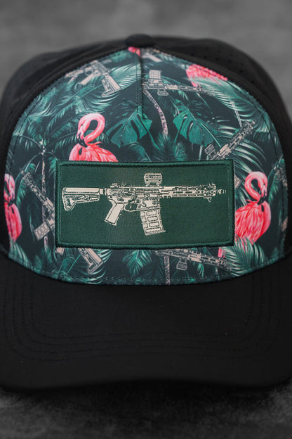 The Loaded Flamingo Hydro Snapback Hat by Tango Charlie Apparel features a black base with green and pink tropical flamingo and rifle patterns, plus a green patch displaying a rifle illustration. The cap is shown resting on a gray surface.