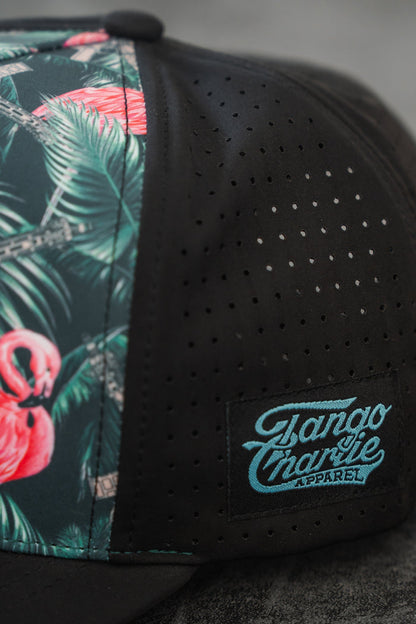 The Loaded Flamingo Hydro Snapback Hat by Tango Charlie Apparel features a black design with a vibrant flamingo and green leaf print on one panel, perforated ventilation holes, and a front Tango Charlie Apparel patch for style and comfort.
