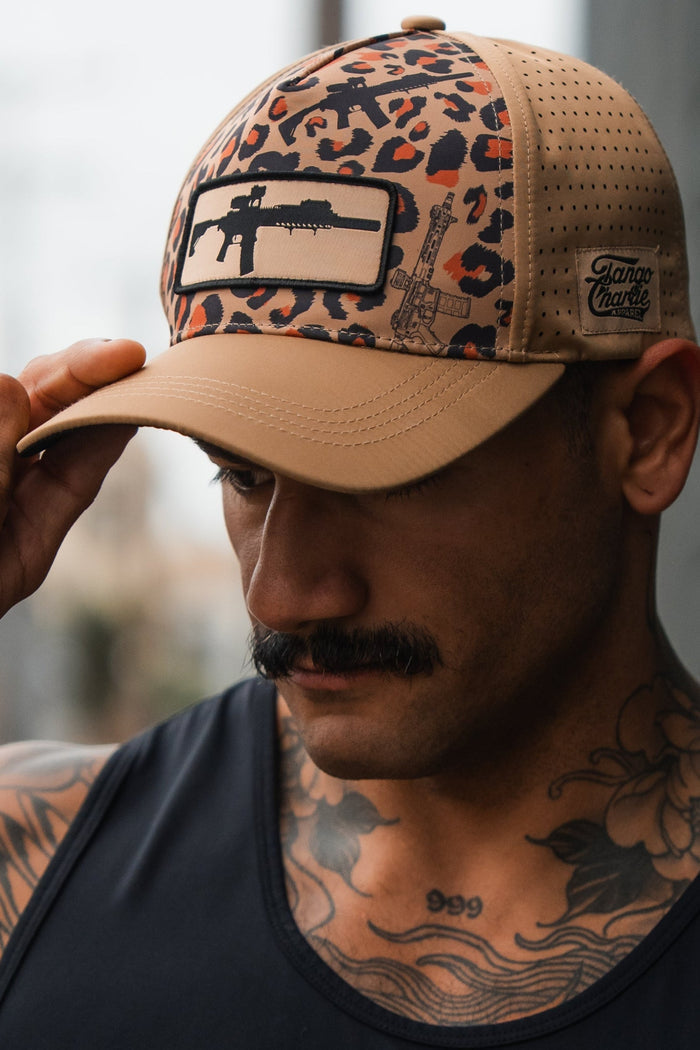A man with a mustache and chest tattoos wears the Loaded Leopard Hydro Snapback Hat by Tango Charlie Apparel, featuring a rifle patch. He looks down, holding the brim of this beige and leopard-print hat, dressed in a black tank top.
