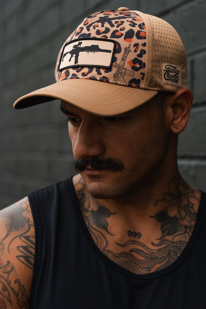 A man with a mustache and tattoos stands against a dark brick wall, looking down. He wears the Tango Charlie Apparel Loaded Leopard Hydro snapback hat with a black rifle patch and a black tank top.