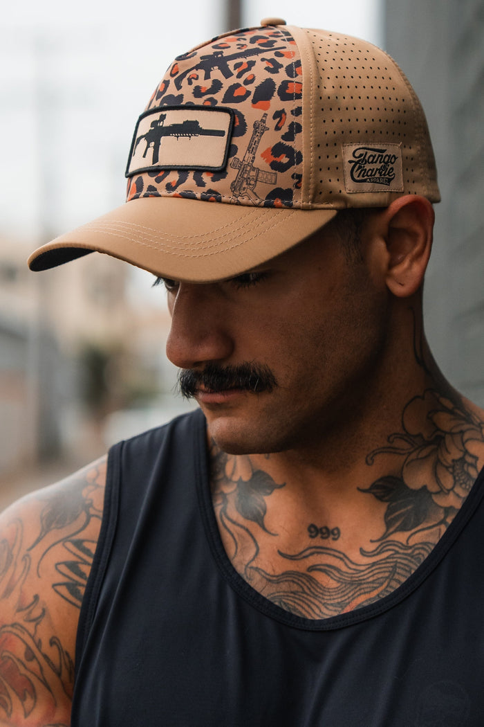 A man with a mustache and floral chest tattoos wears the Tango Charlie Apparel Loaded Leopard Hydro snapback, featuring a tan and leopard print with a rifle design, while his black tank top shows off his tattoos as he looks down.