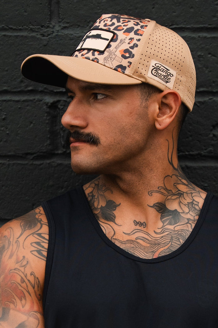 A man with a mustache and chest tattoos, wearing a sleeveless black shirt and the Loaded Leopard Hydro Snapback Hat by Tango Charlie Apparel, stands in front of a black brick wall, looking to the side.