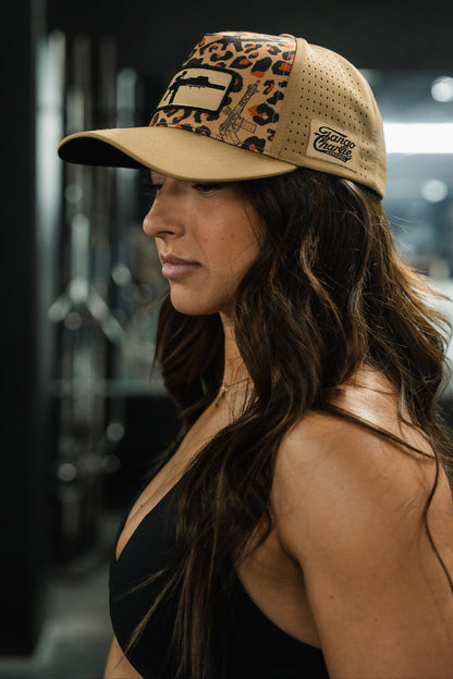 A woman with long brown hair wears the Loaded Leopard Hydro Snapback Hat by Tango Charlie Apparel and a black top, standing indoors with a neutral expression. The background looks like a gym or fitness center.