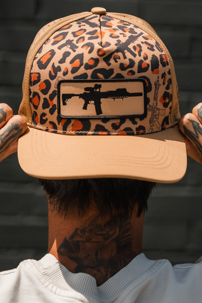 A person faces away from the camera wearing the Loaded Leopard Hydro snapback hat by Tango Charlie Apparel, which features a beige and leopard print design with a black assault rifle patch. The person has finger and neck tattoos.