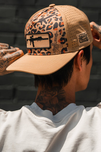 A tattooed person adjusts the Loaded Leopard Hydro Snapback Hat by Tango Charlie Apparel, featuring a gun patch and "Damn Hands" on a beige tag. They're seen from behind in a white shirt against a dark brick wall.