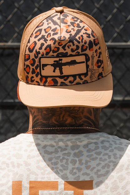A person with neck tattoos stands by a chain-link fence wearing the Tango Charlie Apparel Loaded Leopard Hydro Snapback Hat, featuring tan and leopard print with a black rifle patch, and a light leopard-patterned shirt.