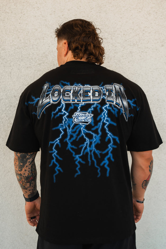 A tattooed individual faces away, wearing Tango Charlie Apparel’s Locked In - Premium Oversized Tee, featuring blue lightning graphics and “LOCKED IN” plus “Stand Chance” text printed on the back.
