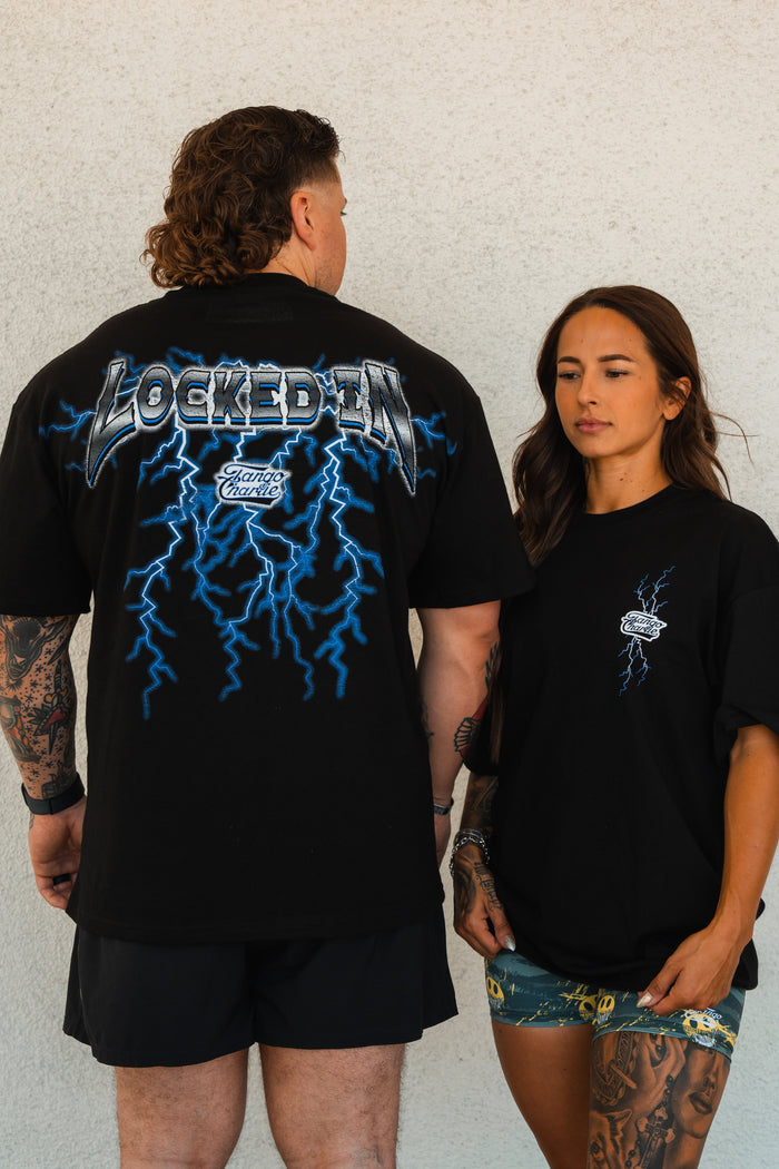 A man shows the back of a Tango Charlie Apparel Locked In - Premium Oversized Tee with blue lightning and LOCKED IN. A woman stands beside him in a comfy unisex tee and patterned shorts, both against a white wall.