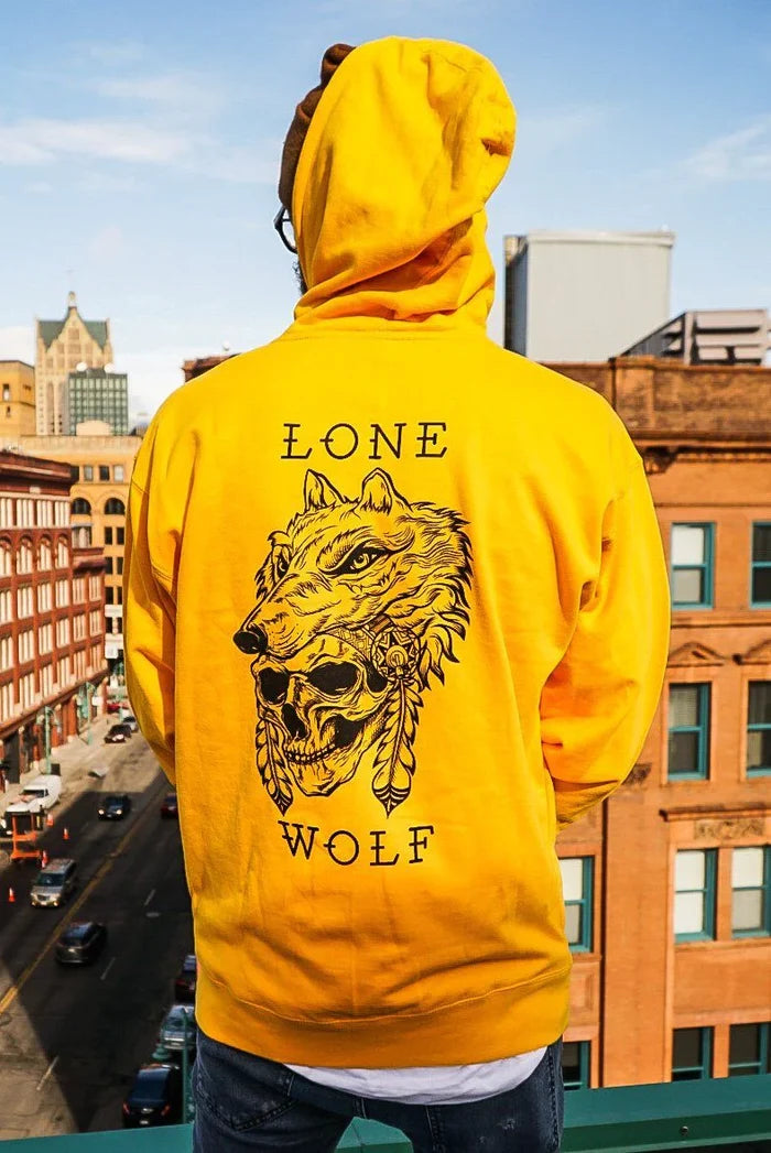 A person in a Relentless Betrayal Lone Wolf GOLD Hoodie stands on a rooftop overlooking city streets. The unisex hoodie showcases a graphic of a wolf with a headdress and the text Lone Wolf, with buildings and clear skies in the background.