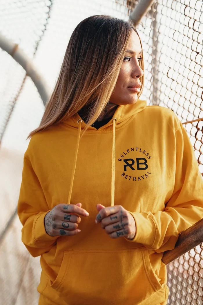 A solitary figure in a Lone Wolf GOLD Hoodie by Relentless Betrayal stands by a chain-link fence, their long hair and tattooed fingers complementing their thoughtful expression, embodying unisex style as they gaze into the distance.
