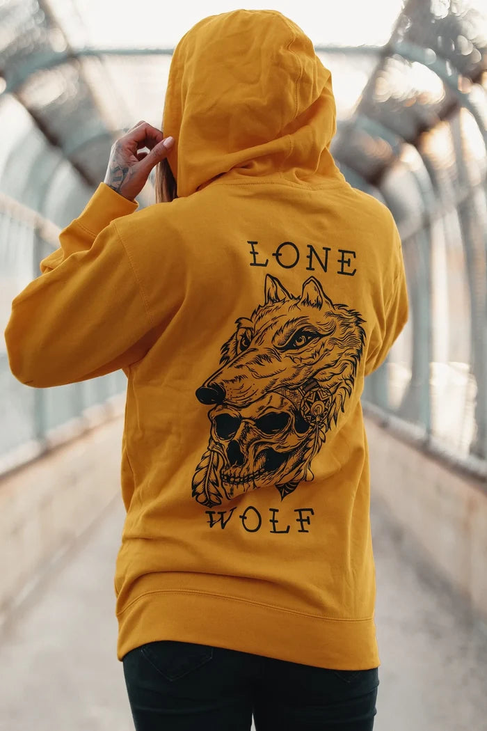 A person in a Lone Wolf GOLD Hoodie by Relentless Betrayal, featuring a wolf illustration and text, stands on a pedestrian bridge with metal railings and a covered structure. The hood is up as they adjust it with one hand.