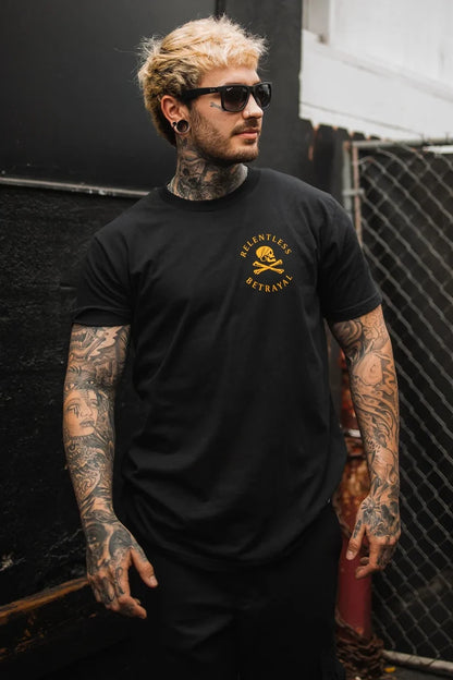A tattooed man in sunglasses stands against a chain-link fence wearing a sleek Relentless Betrayal Lost At Sea Black T-shirt made from fine jersey cotton. The black tee with a yellow design on the left chest highlights his inked arms and neck, all while he maintains a serious expression.