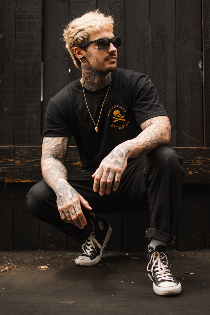 A tattooed man with blond hair and sunglasses crouches in front of a dark wooden fence. He wears a Relentless Betrayal Lost At Sea Black T-shirt made of fine jersey cotton, black pants, black Converse shoes, and a gold chain that enhances the edgy UNISEX style.