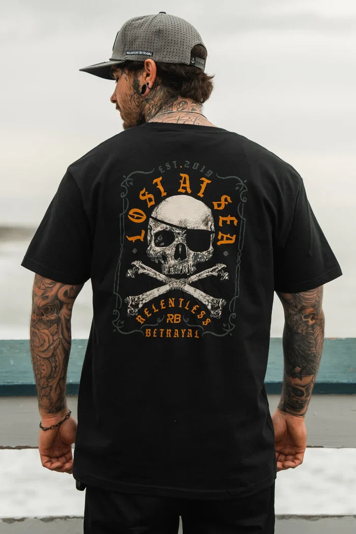 A bearded, tattooed man in a cap stands outdoors by a railing, wearing the black Lost At Sea Crossbones Tee by Relentless Betrayal. The tee showcases a skull and crossbones design with Lost at Sea, Relentless, and Betrayal text. The background is softly blurred as he looks away.