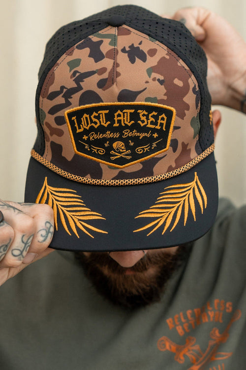 A person adjusts their Lost At Sea Frog Camo snapback by Relentless Betrayal, its patch glinting. The hat, with golden palm leaf embroidery and water resistance, complements their green shirt featuring a matching design.