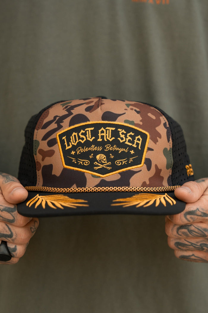 A person holding a water-resistant Lost At Sea Frog Camo Snapback by Relentless Betrayal, featuring gold embroidered skull and crossbones accents, complements their tattoos and green shirt.