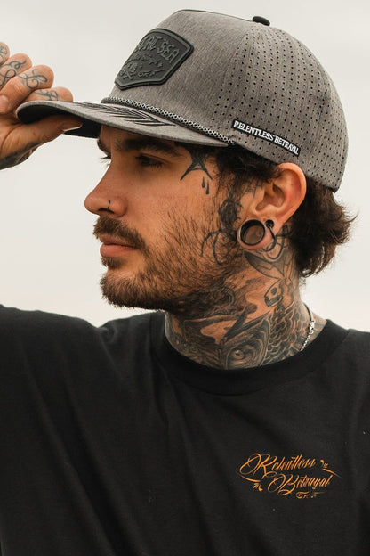 A man with tattoos is wearing a Lost At Sea Gunmetal PVC Snapback by Relentless Betrayal, complementing his large gauged earrings, nose ring, and a black shirt with orange text as he gazes to the right.