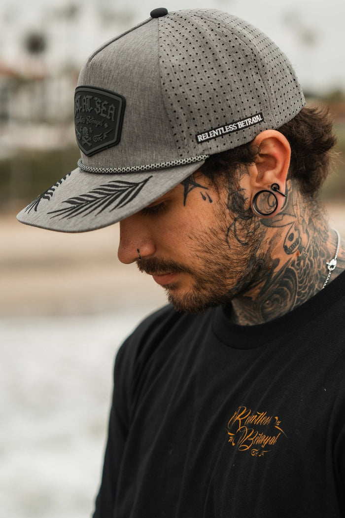 A man with neck tattoos and ear gauges wears the Lost At Sea Gunmetal PVC Snapback by Relentless Betrayal. He has a nose piercing and dons a black shirt with gold writing, enhancing his style as he stands outdoors against a blurred backdrop.