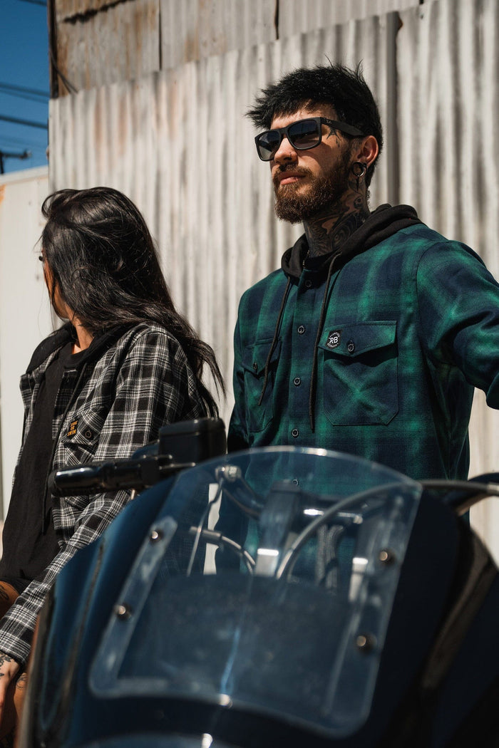 A tattooed man in sunglasses leans on a motorcycle, wearing a Lost At Sea Premium Hooded Flannel from Relentless Betrayal. Beside him, a woman with long dark hair dons another plaid shirt. They pose against a corrugated metal wall, showcasing the rugged charm of ultra-soft flannels.