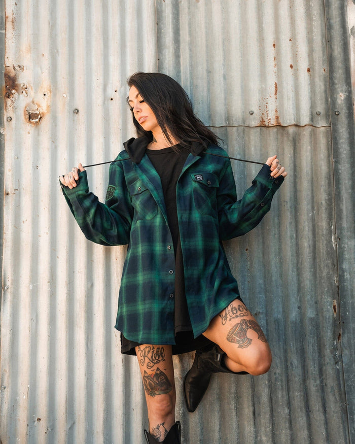 A person with long dark hair, wearing the Relentless Betrayal Lost At Sea Premium Hooded Flannel over a black outfit, stands against a corrugated metal wall. They have leg tattoos and are pulling on the hood strings while resting one foot on the wall in black boots.