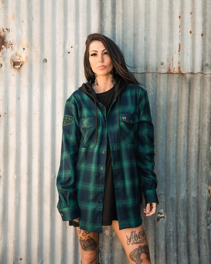 A tattooed person with long hair wears a green and black Lost At Sea Premium Hooded Flannel by Relentless Betrayal over a black hoodie, standing against a corrugated metal wall while looking at the camera.